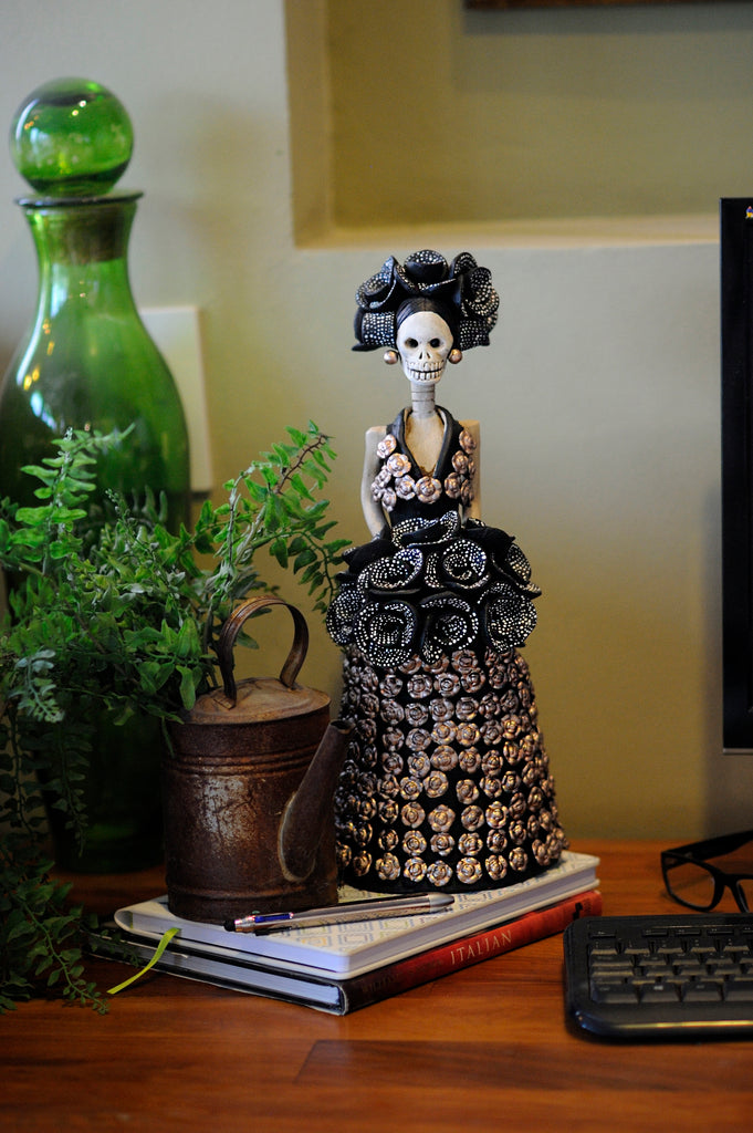 Hand-made clay figurine, Carmina is wearing a black dress, with silver flower accents all over her dress. To accessorize she is holding a black rose bouquet with a white dotted pattern and the same can be seen on her headpiece. All our Catrinas are made in Mexico and shipped worldwide.  