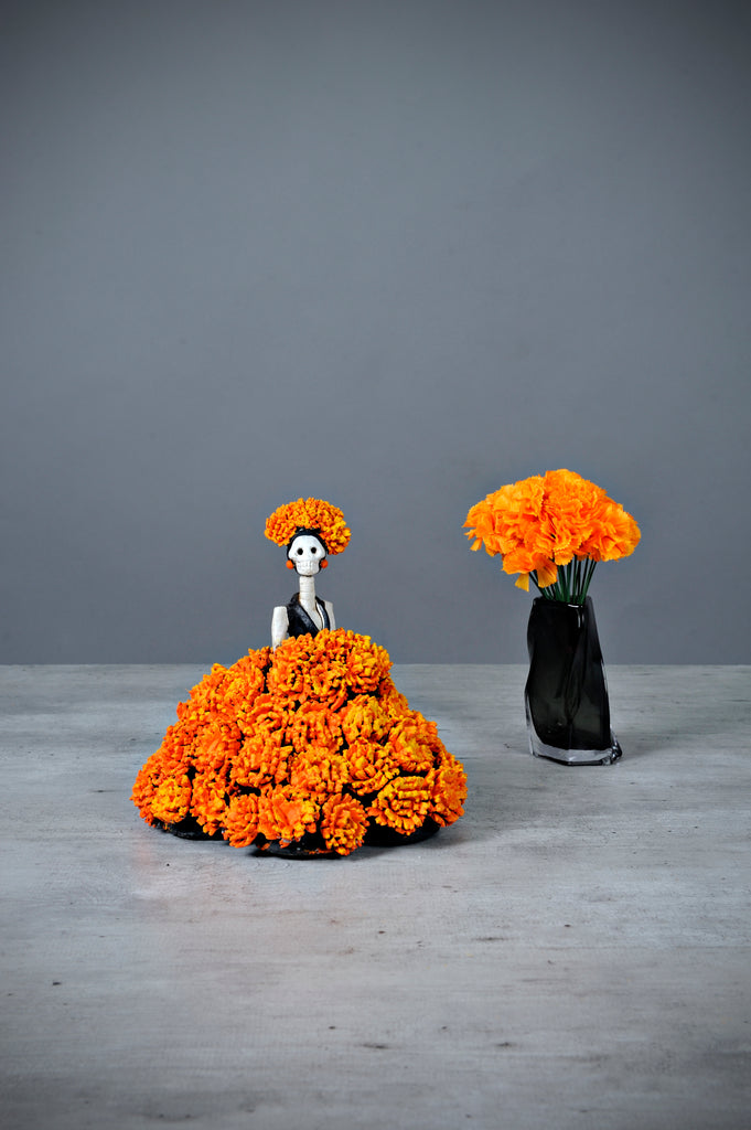 Hand-made clay figurine.Catrina Gisela is a unique Catrina, wearing a stunning ball gown covered in cempasúchil (Marigold) flowers. This type of flower is an important piece used to decorate Day of the Dead altars.  Each flower is hand-made and painted before carefully placing it on her dress. Gisela is the perfect addition to your Day of the Dead altar. All of our Catrinas are made in Mexico and shipped worldwide. Add a classy touch of Mexican art to your home.