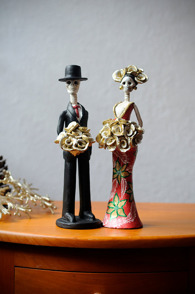 Hand-made clay figurine, Catrina Cynthia is wearing a curve-skimming red dress with gold details on her dress as well as hand-painted poinsettias. She is holding a gold rose bouquet. Her date Diego is also holding a gold rose bouquet. All our Catrinas are made in Mexico and shipped worldwide. Add a classy touch of Mexican culture to your home. 