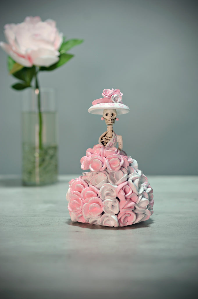 Hand-made clay figurine, Catrina Rosalinda is a small catrina, her dress is covered in pink and white roses, she is wearing a cute white hat with roses that match her dress and bouquet. All our Catrinas are made in Mexico and we ship worldwide. Add a touch of Mexican art to your home. 