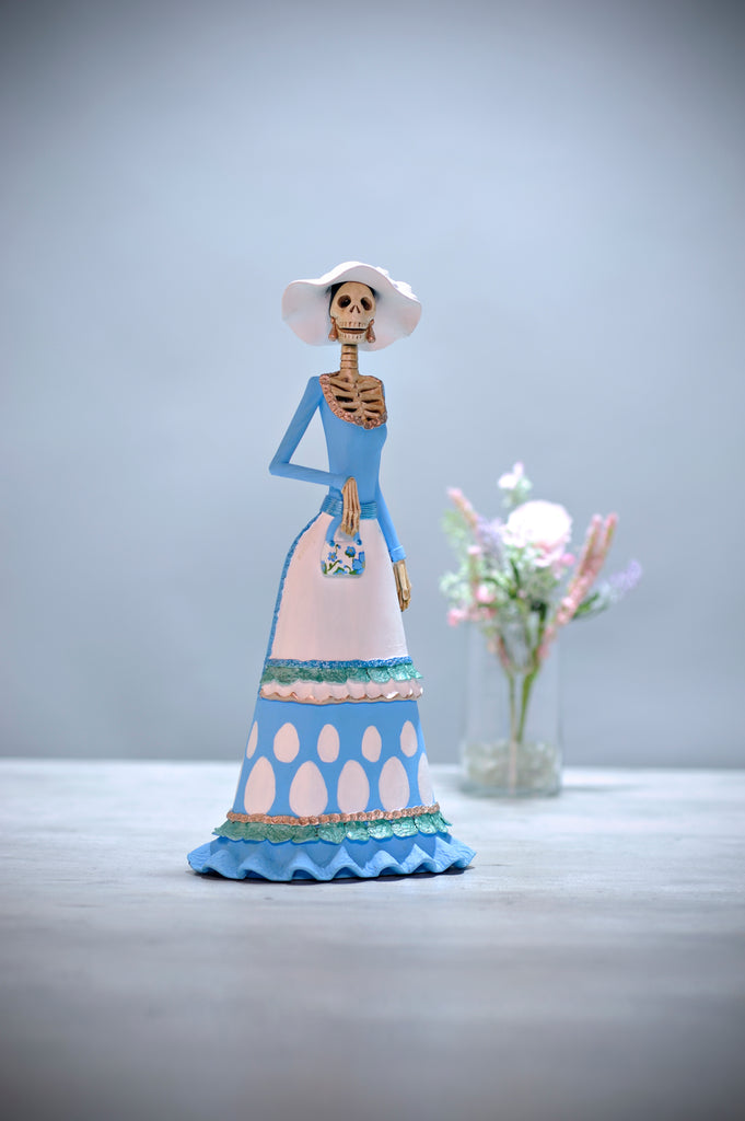 Hand-made clay figurine, Catrina Adela is a beautiful lady wearing a pastel blue dress with exquisite detailing at the base of the dress. To accentuate her figure, she is wearing a white apron that also has ruffles at the bottom. To accessorize her outfit, she opted for a white sunhat and the best part is her purse. The artist painted an incredible pattern onto the purse, making it a unique piece. All our Catrinas are made in Mexico and we ship worldwide. Add a classy touch of Mexican art to your home.