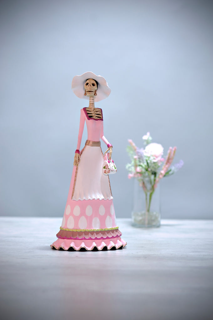 Hand-made clay figurine, Catrina Carmen is part of our Easter collection. This beautiful lady is wearing a pink dress with creative detailing at the base. To accentuate her figure, she is wearing a white apron that matches her hat, and the most impressive part is her purse. The artist painted a stunning pattern onto the purse making it a unique piece. All our Catrinas are made in Mexico and we ship worldwide. Add a classy touch of Mexican culture to your home.