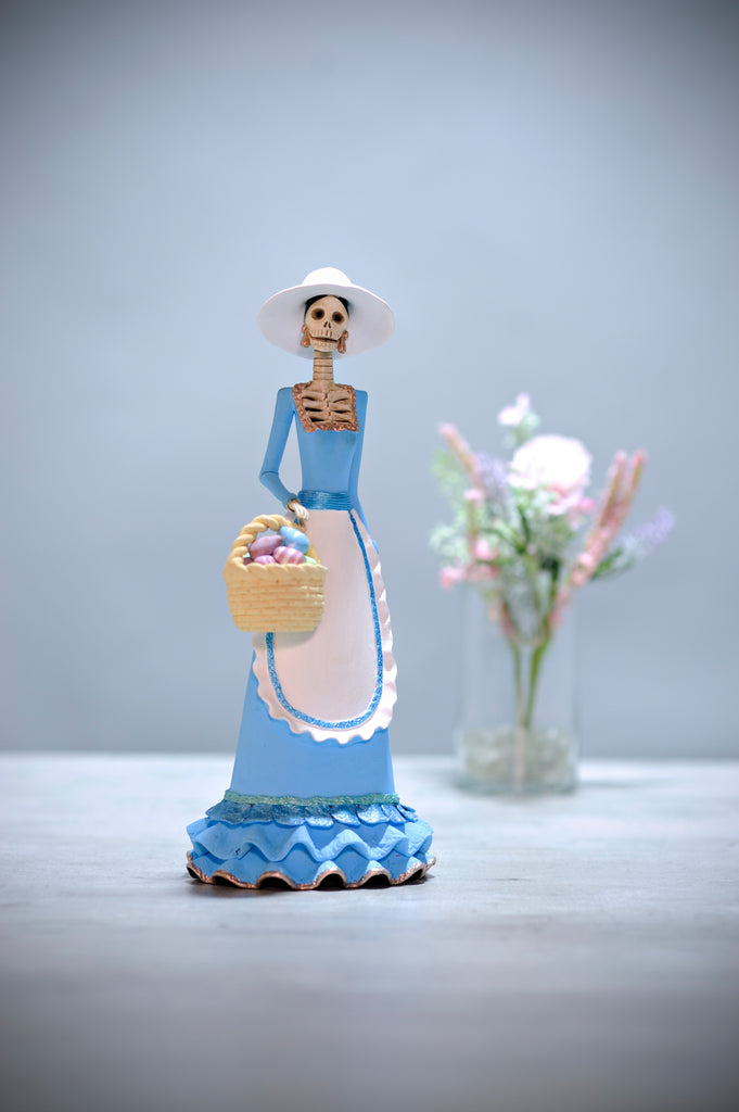 Hand-made clay figurine, Catrina Dora is part of our Easter collection. She is wearing a beautiful pastel blue dress with a teardrop shaped apron in white. She is also wearing a hat that matches her apron. She is holding an Easter egg basket filled with colorful hand-painted eggs. All our Catrinas are made in Mexico and we ship worldwide. Add a classy touch of Mexican culture to your home.