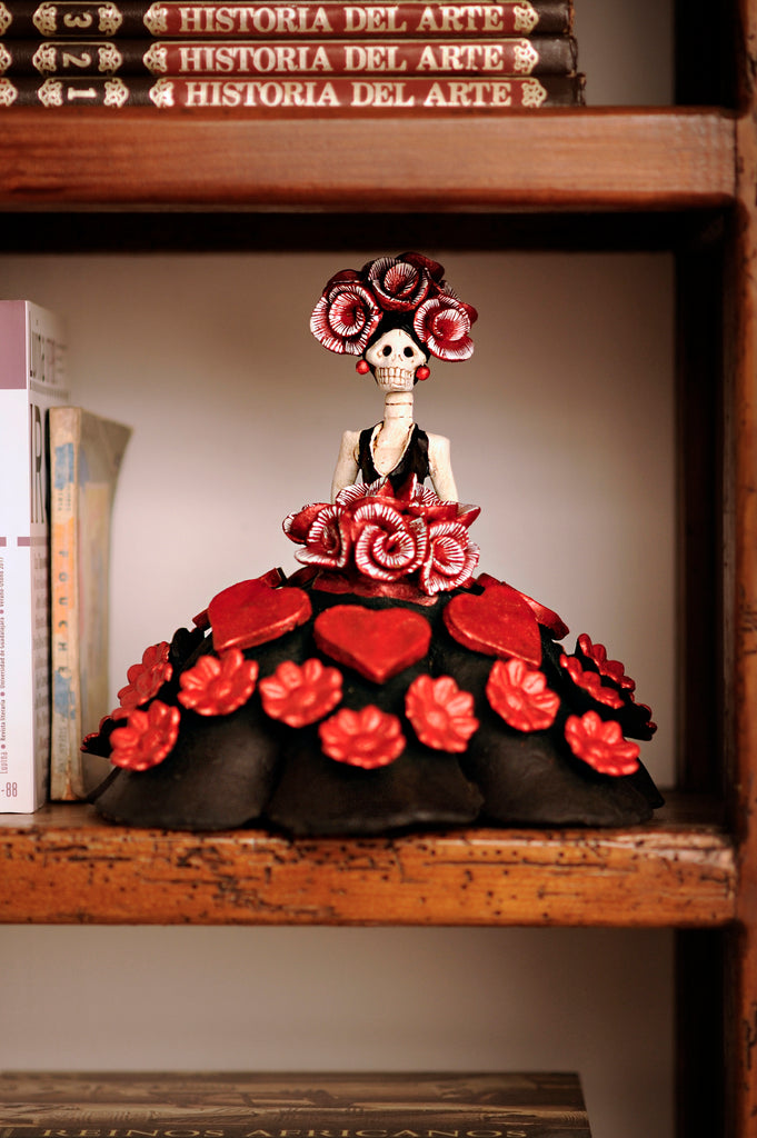 Catrina Carlota is a hand-made clay figurine, Carlota is wearing a black ball gown with red hearts and red flowers scattered around her dress, she is also carrying a red rose bouquet that matches the flowers on her headpiece. All our Catrinas are made in Mexico and we can ship worldwide. Add a classy touch of Mexican art to your home.
