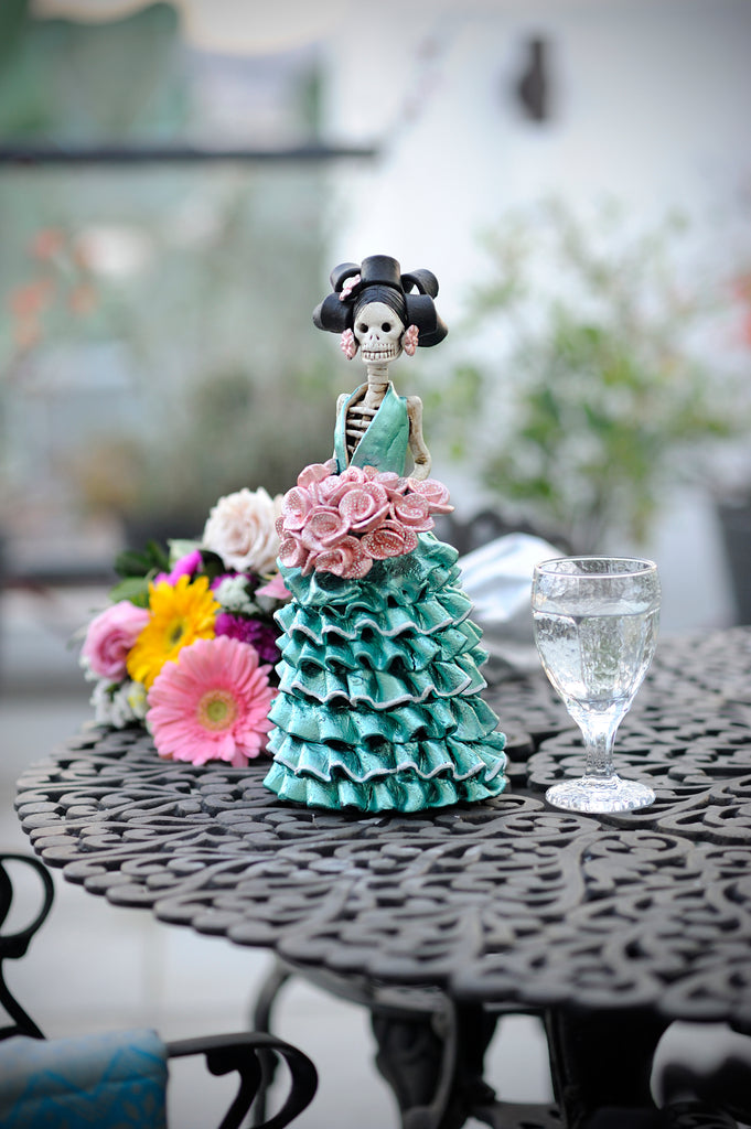 Hand-made clay figurine, wearing a turquoise ruffled dress. Berenice is holding a beautiful pink dotted rose bouquet. Add a classy touch of Mexican art to your home or office. All our Catrinas are made in Mexico. We ship worldwide.