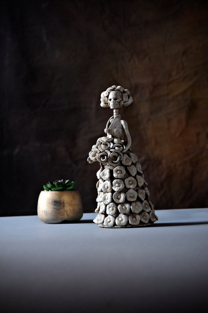 Hand-made clay figurine, painted in ivory color, this catrina has flowers placed all over her dress and she is holding a rose bouquet. All our Catrinas are made in Mexico, we ship worldwide.