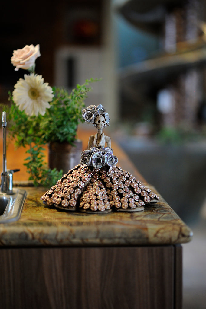 Catrina Marissa is a small hand-made clay figurine. Her gown is decorated with small flowers and she is holding a silver rose bouquet with a black dotted pattern. Her headpiece is made up of the same flowers as her bouquet. This catrina is a perfect gift for Mother's Day. All our Catrinas are made in Mexico and we can ship worldwide. Add a classy touch of Mexican culture to your home.
