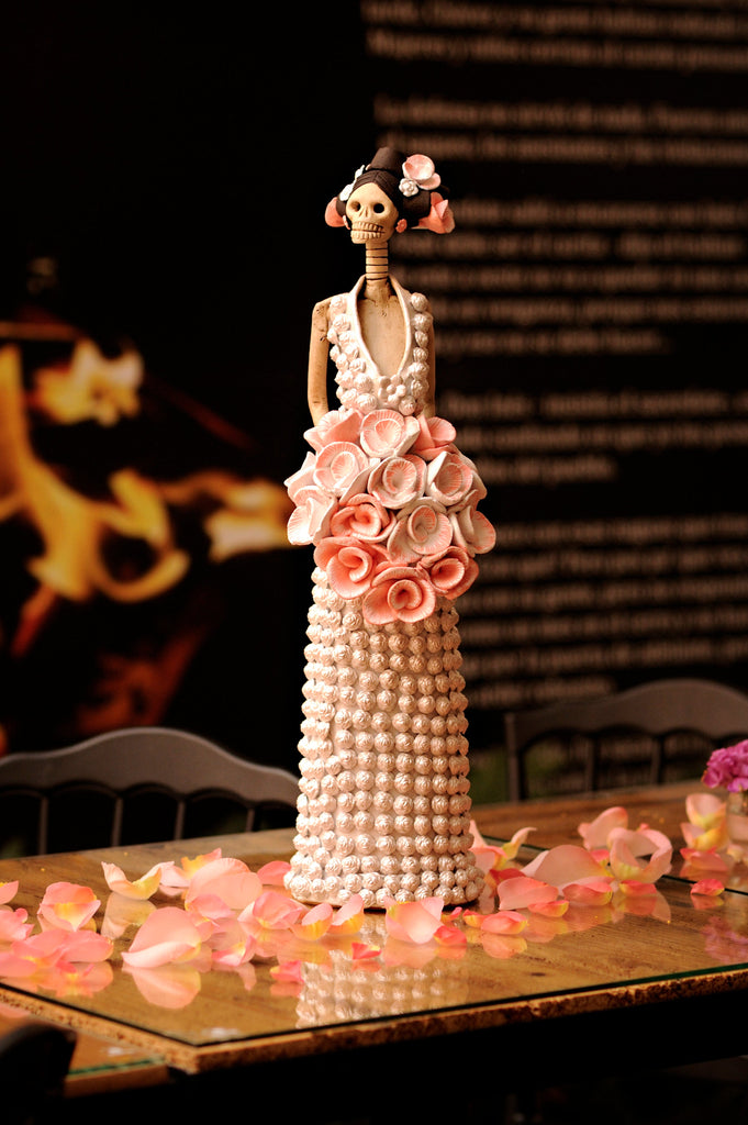 Hand-made clay figurine, Catrina Daniela is a beautiful bride wearing a white dress covered in little roses. She is holding a bouquet of roses that are painted in light pink and white. These roses match her beautiful headpiece. All our Catrinas are made in Mexico and we ship worldwide. Add a classy touch of Mexican art your home.