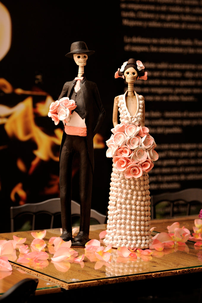 Hand-made clay figurines, Daniela and Brandon are a Catrina couple who are celebrating their recent marriage. She is wearing a beautiful white dress covered in little white rosebuds, while her husband is wearing a classic tux with a pink cummerbund that matches the rose bouquets they are both holding. All our Catrinas are made in Mexico and we ship worldwide. Add a touch of classy Mexican art to your home.