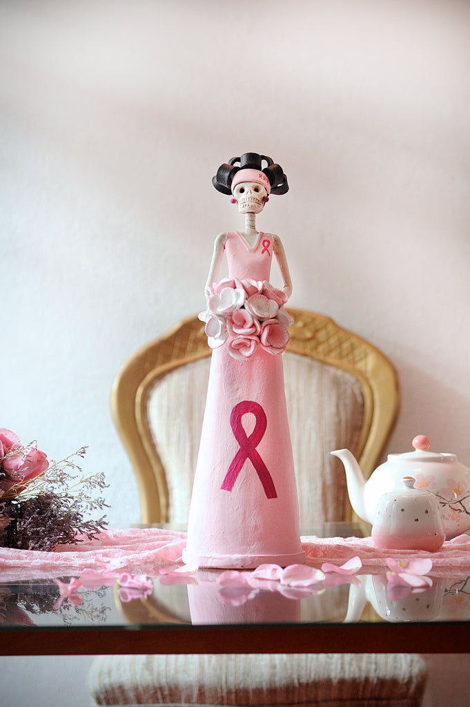 Hand-made clay figurine, Catrina Cecilia is wearing a light pink dress with a pink ribbon on the skirt, in support of Breast Cancer Awareness. All our Catrinas are made in Mexico and we ship worldwide. Add a classy touch of Mexican culture to your home. 