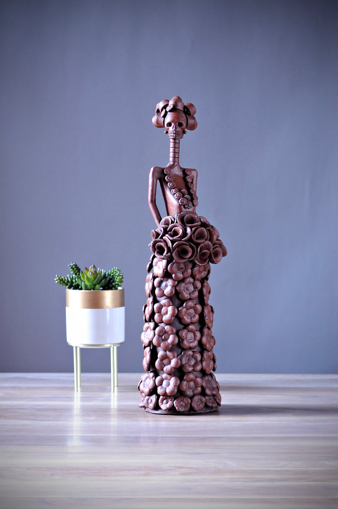 Hand-made clay figurine, Catrina Natalia is a beautiful Catrina wearing a long A-line shaped dress with flowers all around her dress. She is also holding a rose bouquet. Add a touch of Mexican art to your home. All our Catrinas are made in Mexico and ship worldwide.  